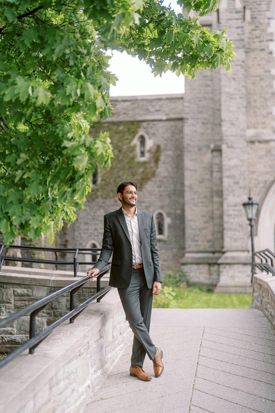 A man in a suit leans casually against a stone railing with historic university architecture and green trees in the background.