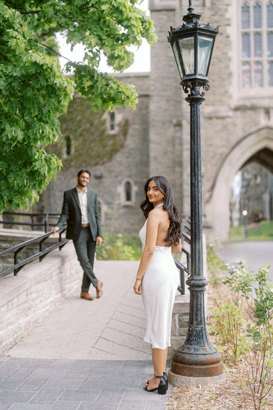 Tej and Priyana stand on a stone pathway at the University of Toronto, with Priyana in the foreground and Tej leaning on a railing in the background.