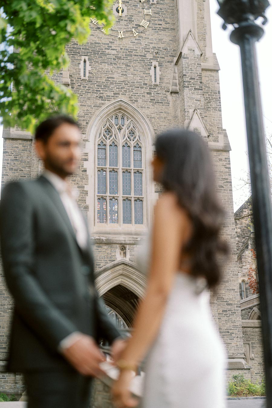 A couple stands hand in hand in front of a historic stone clock tower at the University of Toronto St. George campus.