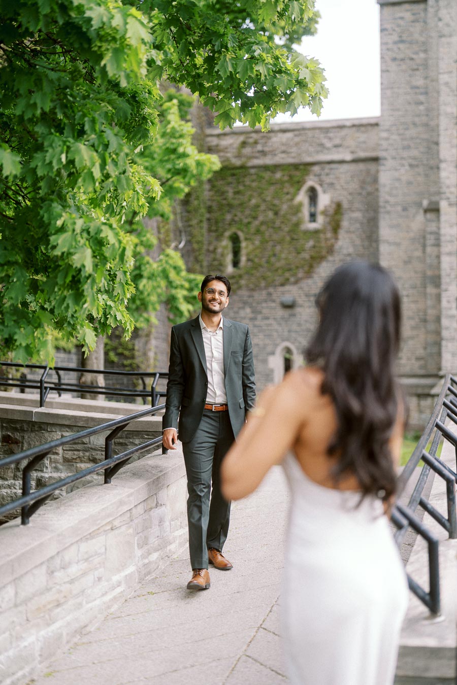 Tej walks toward Priyana on a stone pathway at the University of Toronto campus, with leafy green trees and historic stone buildings in the background.
