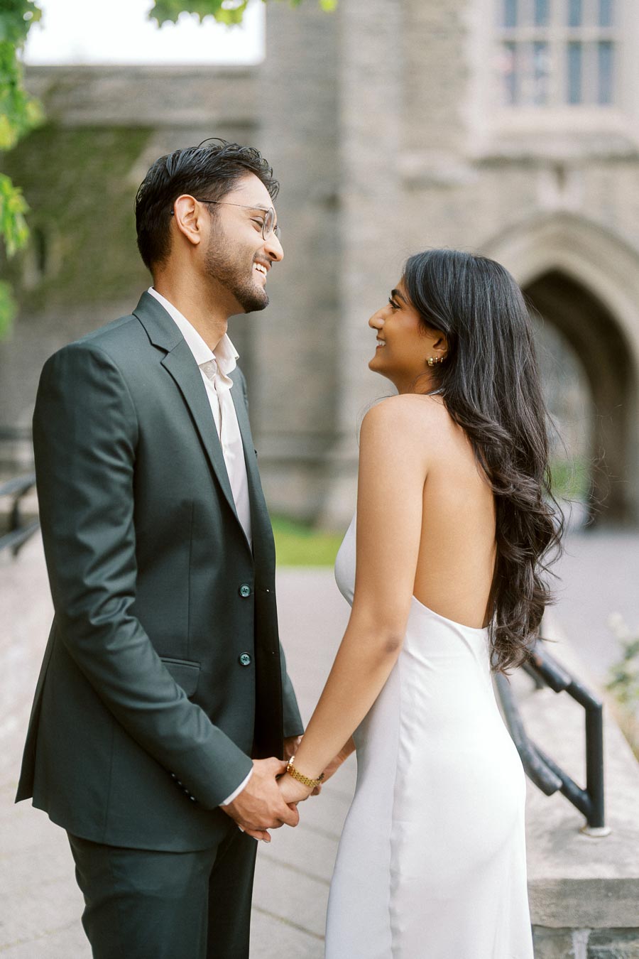 A couple stands close together holding hands and smiling at each other, with stone university architecture in the background at the University of Toronto.