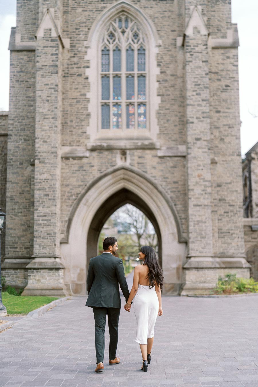 A couple holding hands and walking away toward a large stone archway at the University of Toronto campus.
