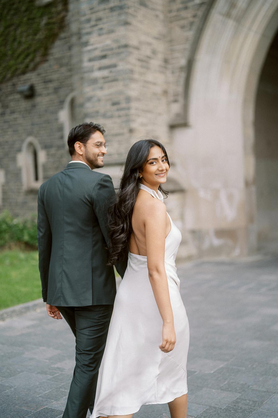 Tej and Priyana walk together in formal attire near a historic stone archway at the University of Toronto St. George campus.