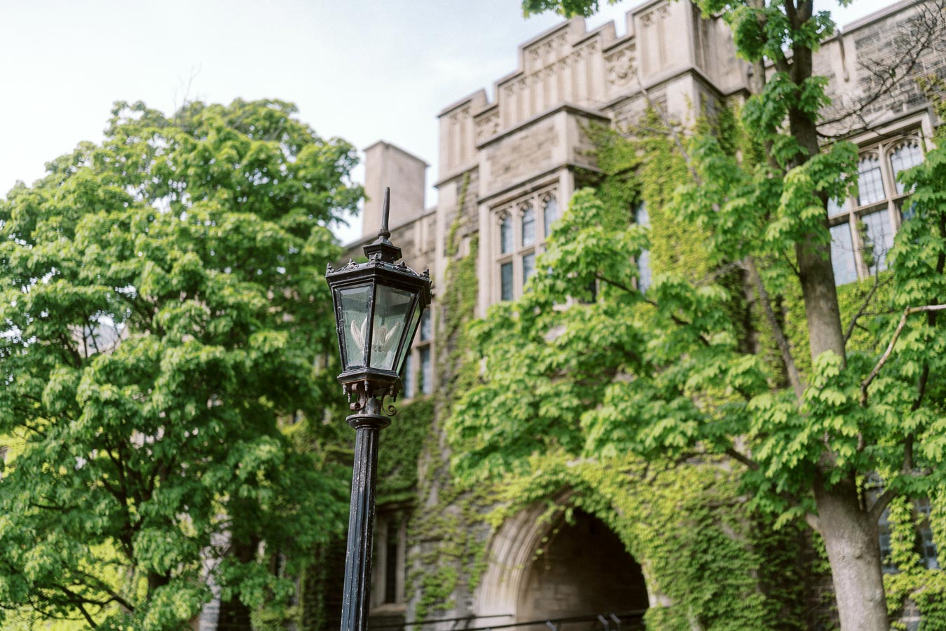 A vintage black lamp post in front of an ivy-covered stone building with arched windows at the University of Toronto St. George campus.