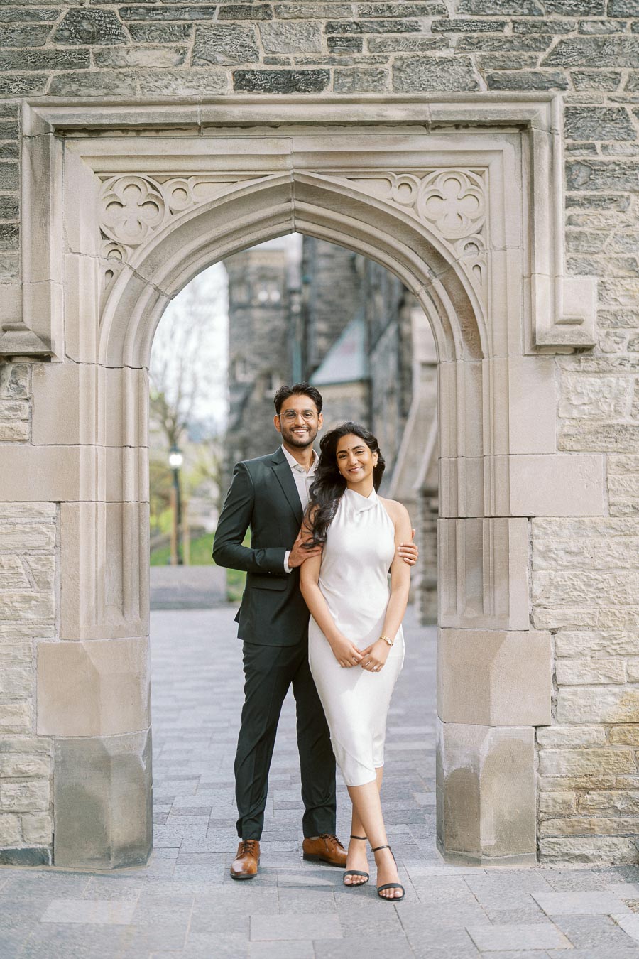 A couple stands close together beneath a detailed stone archway on the University of Toronto campus, with historic architecture in the background.