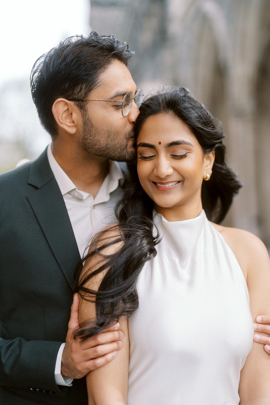 Tej gently kisses Priyana on the forehead while holding her, with blurred stone arches in the background at University of Toronto.
