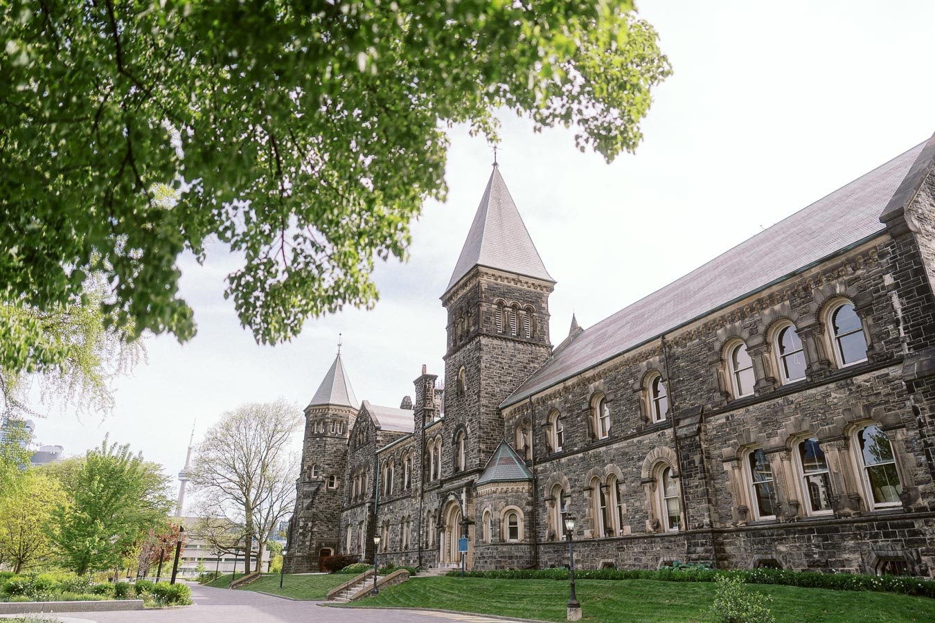 A historic stone building with arched windows and towers at the University of Toronto St. George campus, surrounded by green trees and grass.