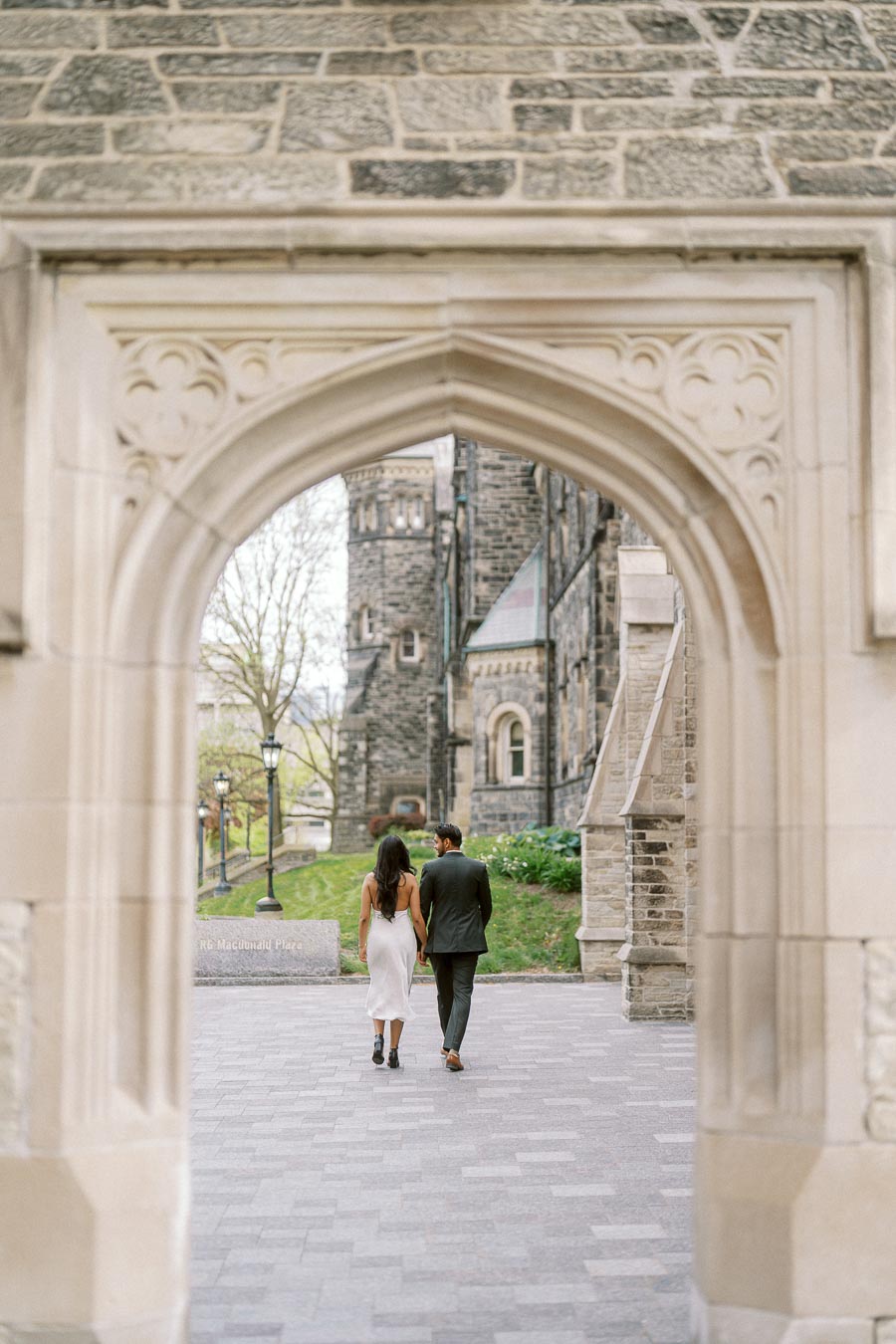 A couple walks hand in hand away from the camera through a large stone archway at the University of Toronto St. George campus.