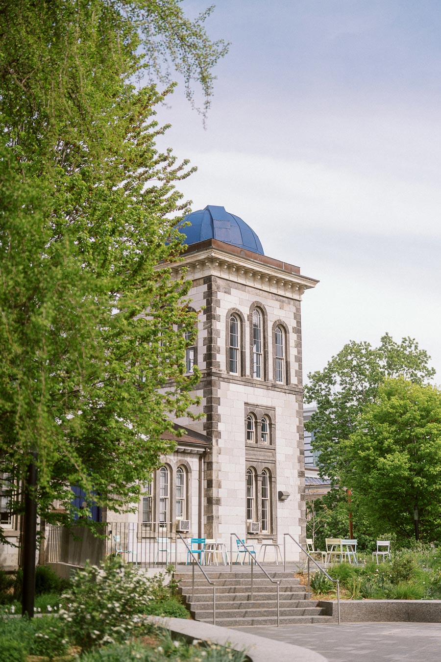 A historic stone building with a blue dome at the University of Toronto is framed by green trees and outdoor tables in spring.
