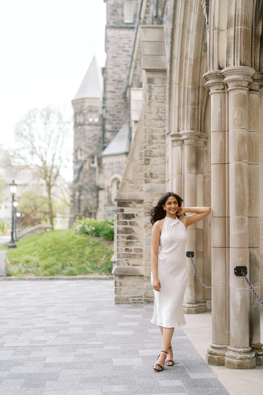A woman in a white dress poses by a stone column under a historic archway at the University of Toronto campus.
