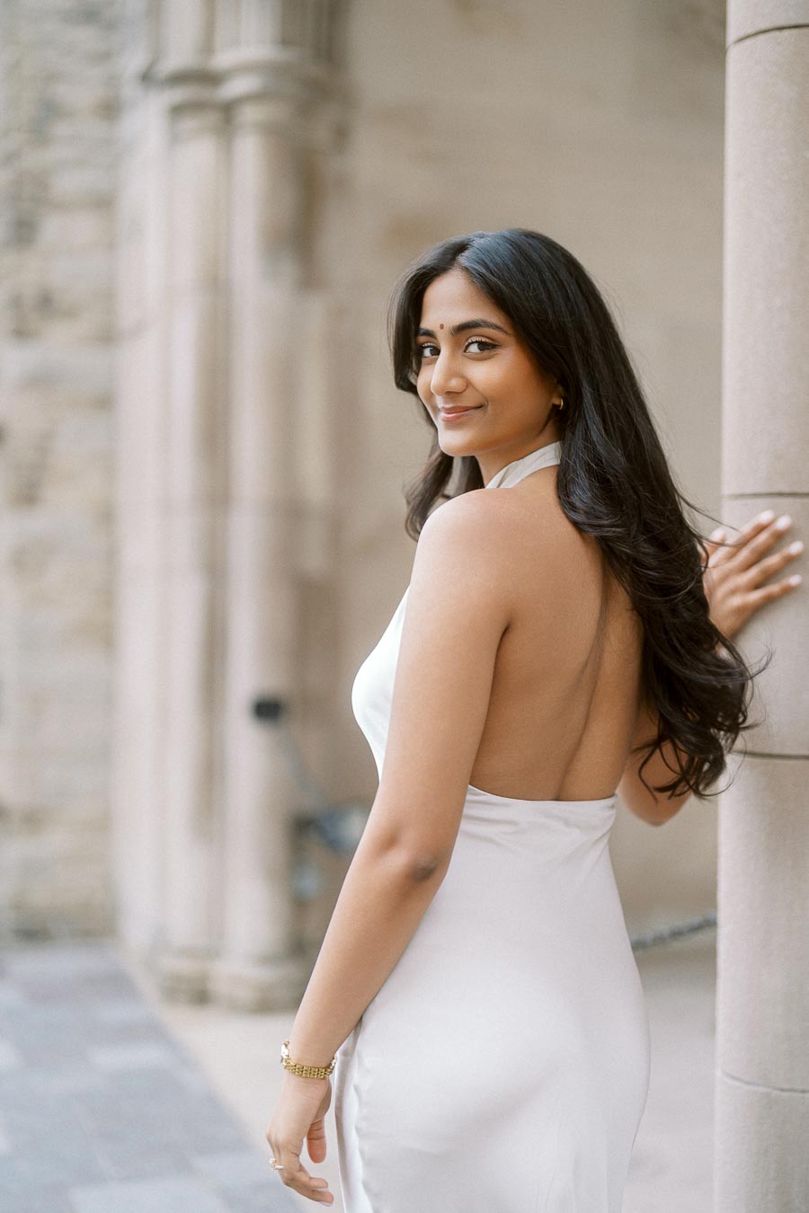 Priyana in a white dress stands by a stone column at the University of Toronto, looking over her shoulder with a soft smile.