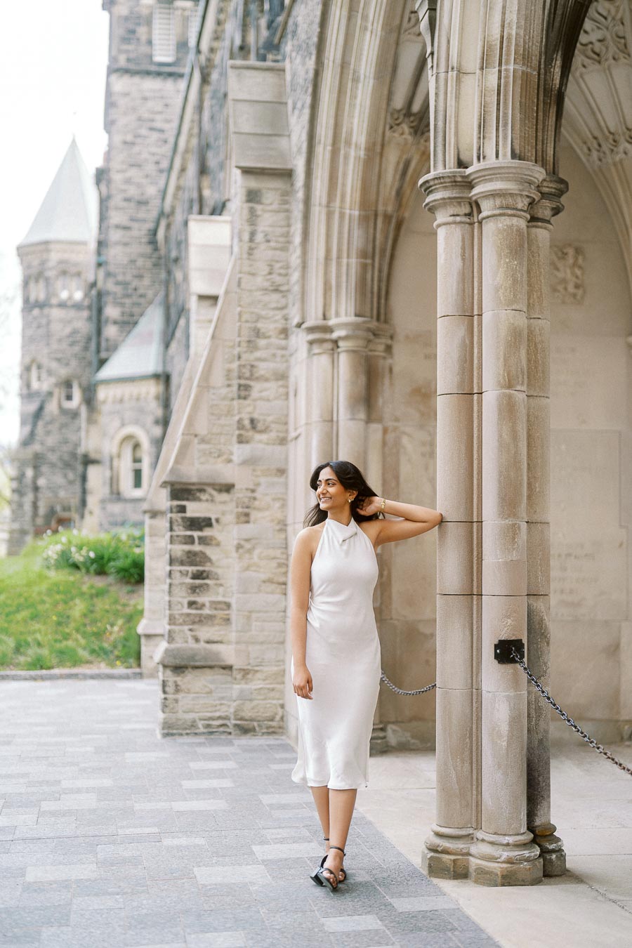 A woman in a white dress stands under a stone archway at the University of Toronto St. George campus, with historic architecture in the background.