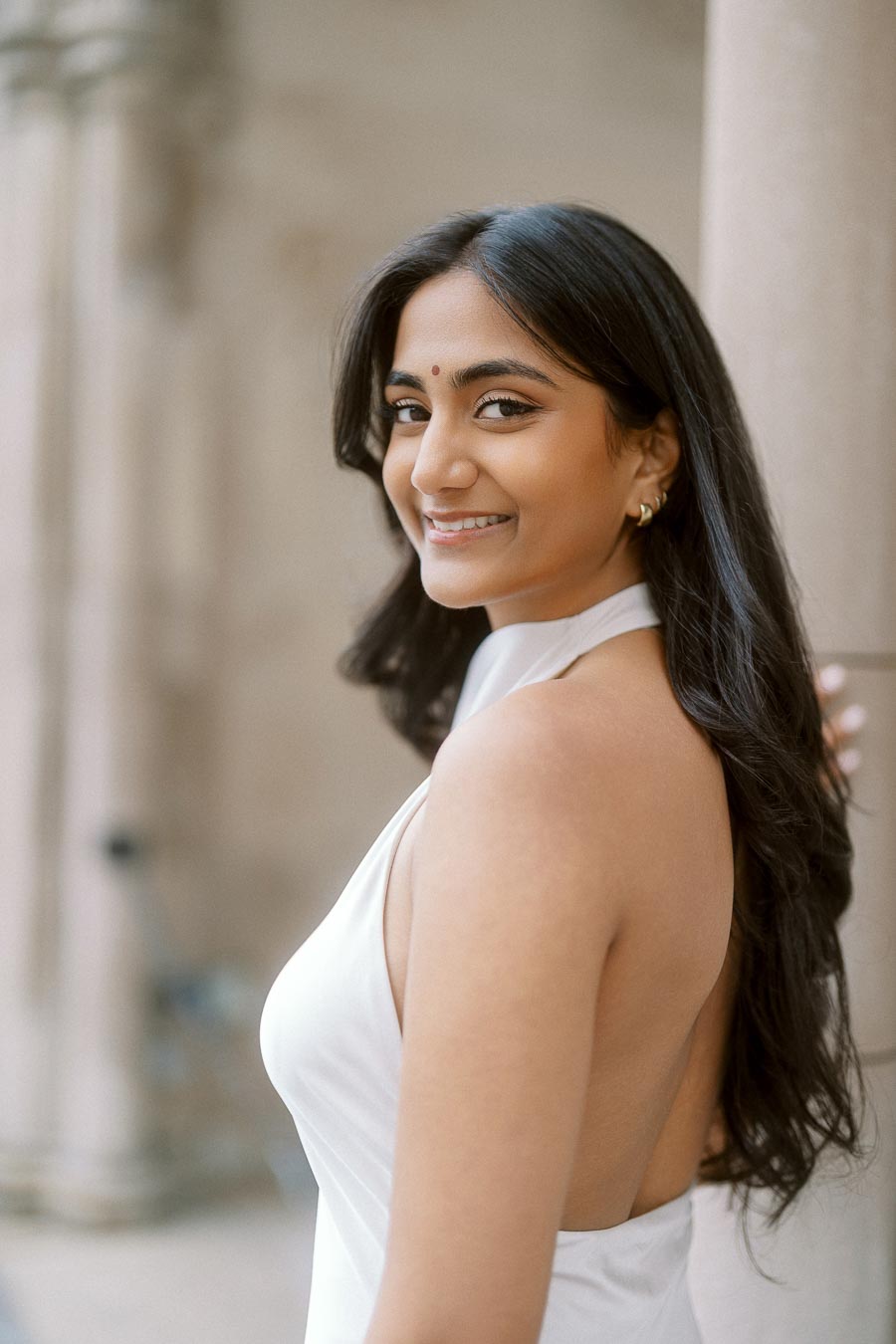 A woman with long dark hair in a white dress smiles at the camera, standing beside a stone column at the University of Toronto.