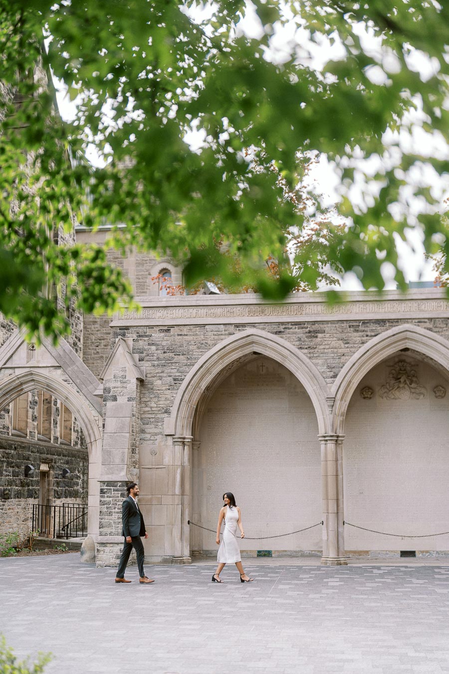 A couple walks side by side in front of large stone archways at the University of Toronto, with green tree branches overhead.