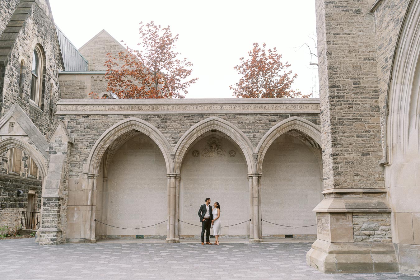 A couple stands closely together under a large stone archway at the University of Toronto, with historic architecture and trees in the background.