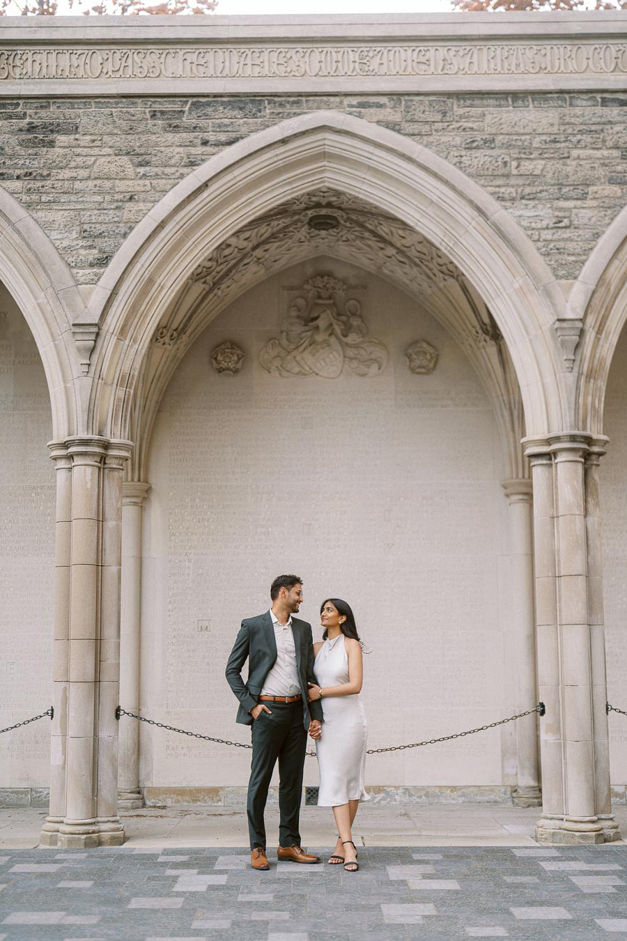 Tej and Priyana stand side by side under a large Gothic stone archway on the University of Toronto campus.