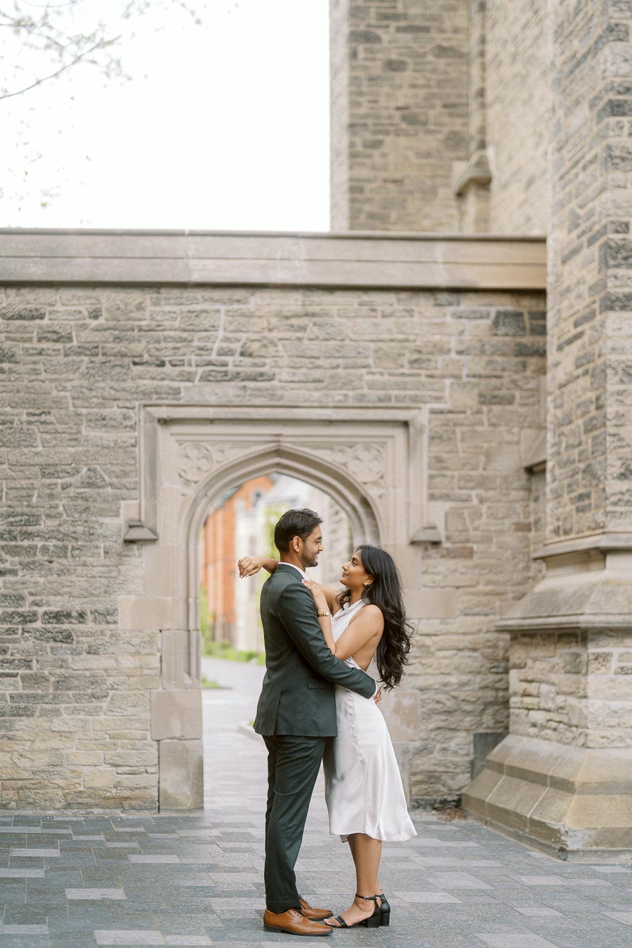 A couple stands close together, gazing at each other in front of a historic stone archway at the University of Toronto.