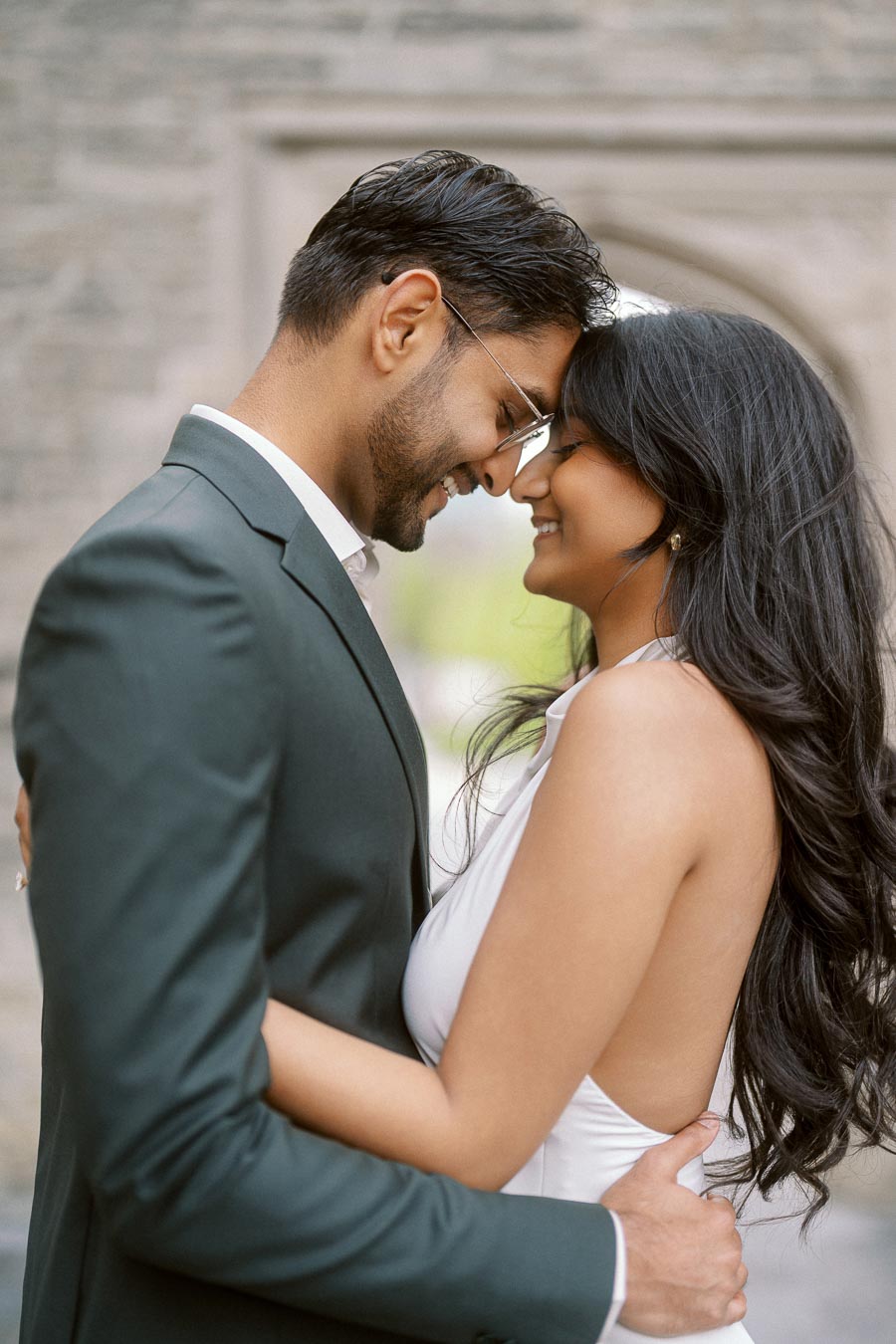 A couple stands closely together, smiling with their foreheads touching, in front of a stone archway at University of Toronto.