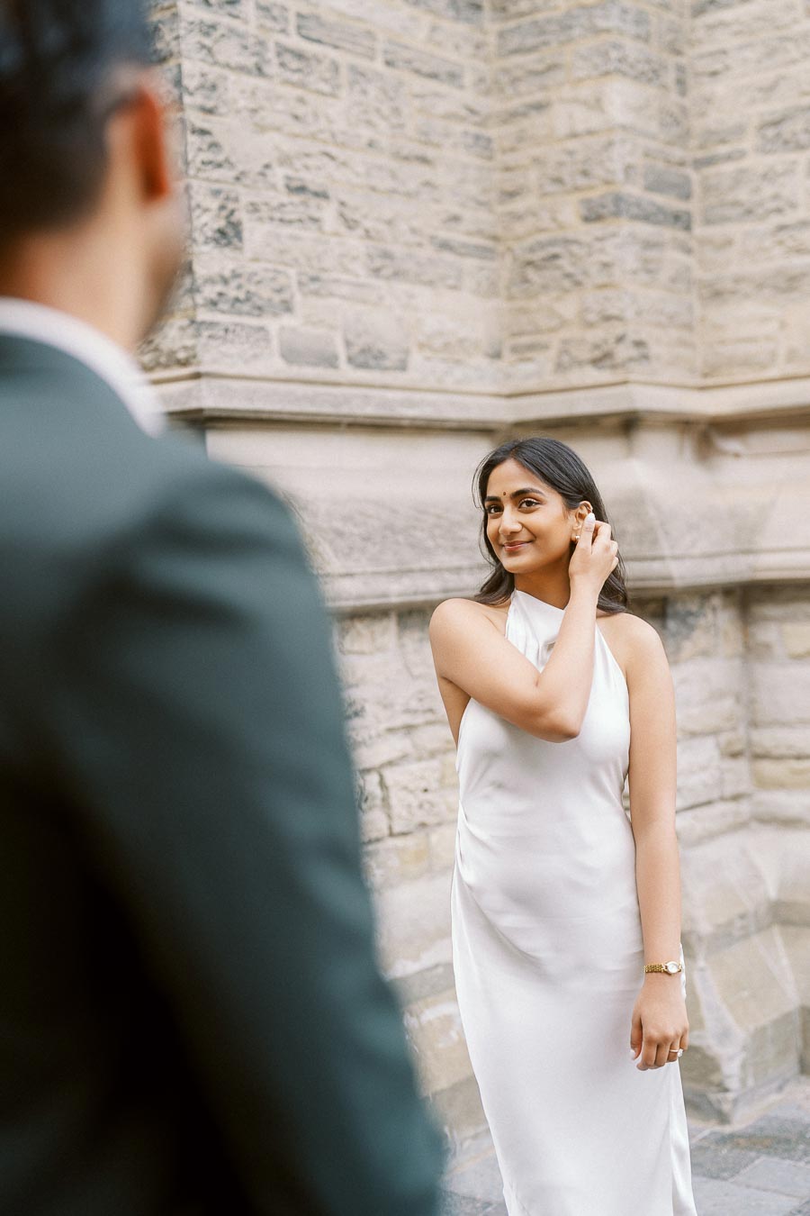 Priyana stands in a white dress by a stone wall at University of Toronto while Tej is partially visible in the foreground.