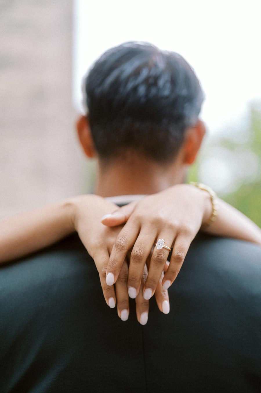 Close-up of a woman's hands with an engagement ring resting on a man's shoulders outdoors at the University of Toronto.