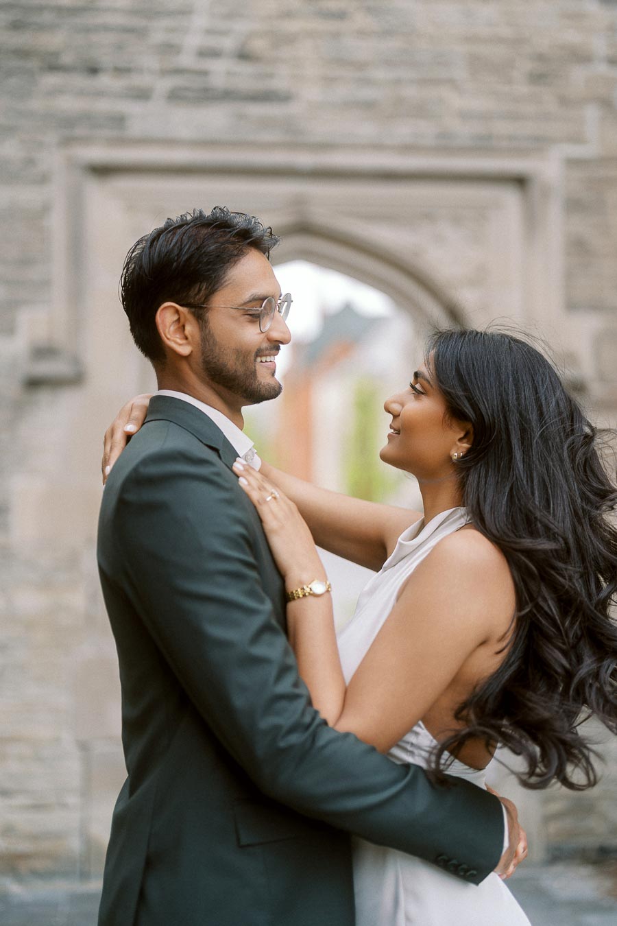 Tej and Priyana stand closely together, smiling and holding each other in front of a stone archway at University of Toronto.