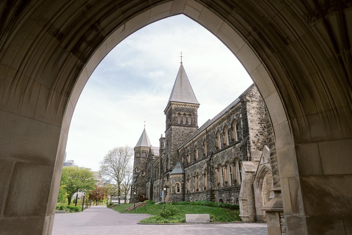 Stone university building with pointed towers seen through a large stone archway at the University of Toronto St. George campus.