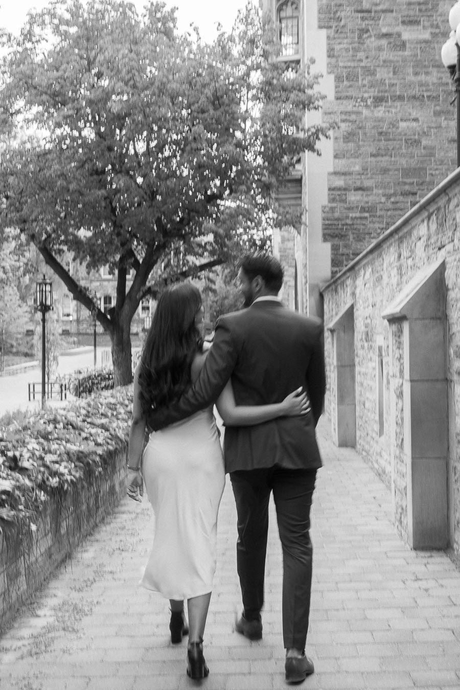 A couple walks arm in arm along a stone pathway beside ivy-covered walls at the University of Toronto campus.
