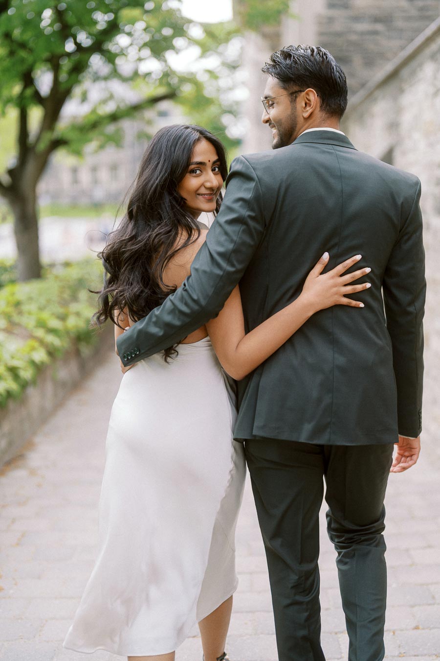Tej and Priyana walk closely together on a tree-lined stone path at the University of Toronto campus, looking back over their shoulders.