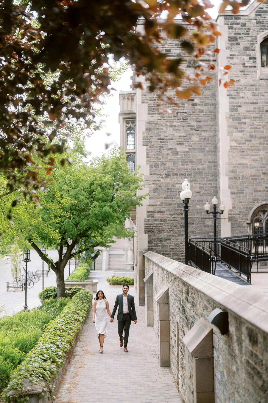 A couple walks hand in hand down a stone path surrounded by greenery and historic stone buildings at the University of Toronto.