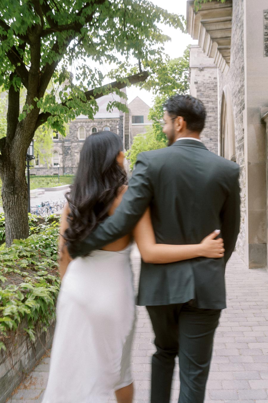 Tej and Priyana walk arm in arm along a tree-lined path beside historic stone buildings at the University of Toronto St. George campus.