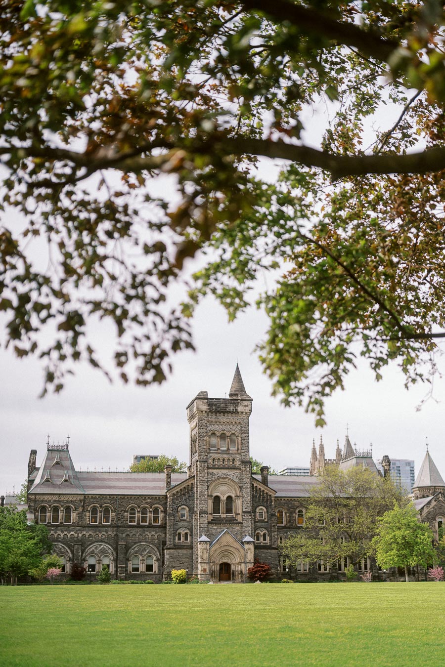 A historic stone building with a central tower at the University of Toronto, seen across a green lawn and framed by leafy tree branches.
