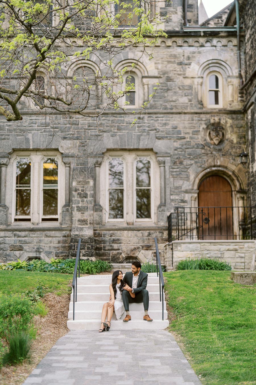 A couple sits closely together on outdoor stone steps in front of a historic stone building at the University of Toronto St. George campus, surrounded by green grass and budding trees.