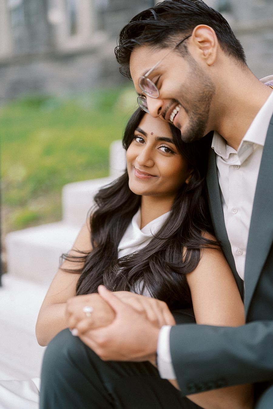 Tej and Priyana sit together on outdoor steps, holding hands, with Priyana's engagement ring visible at the University of Toronto.