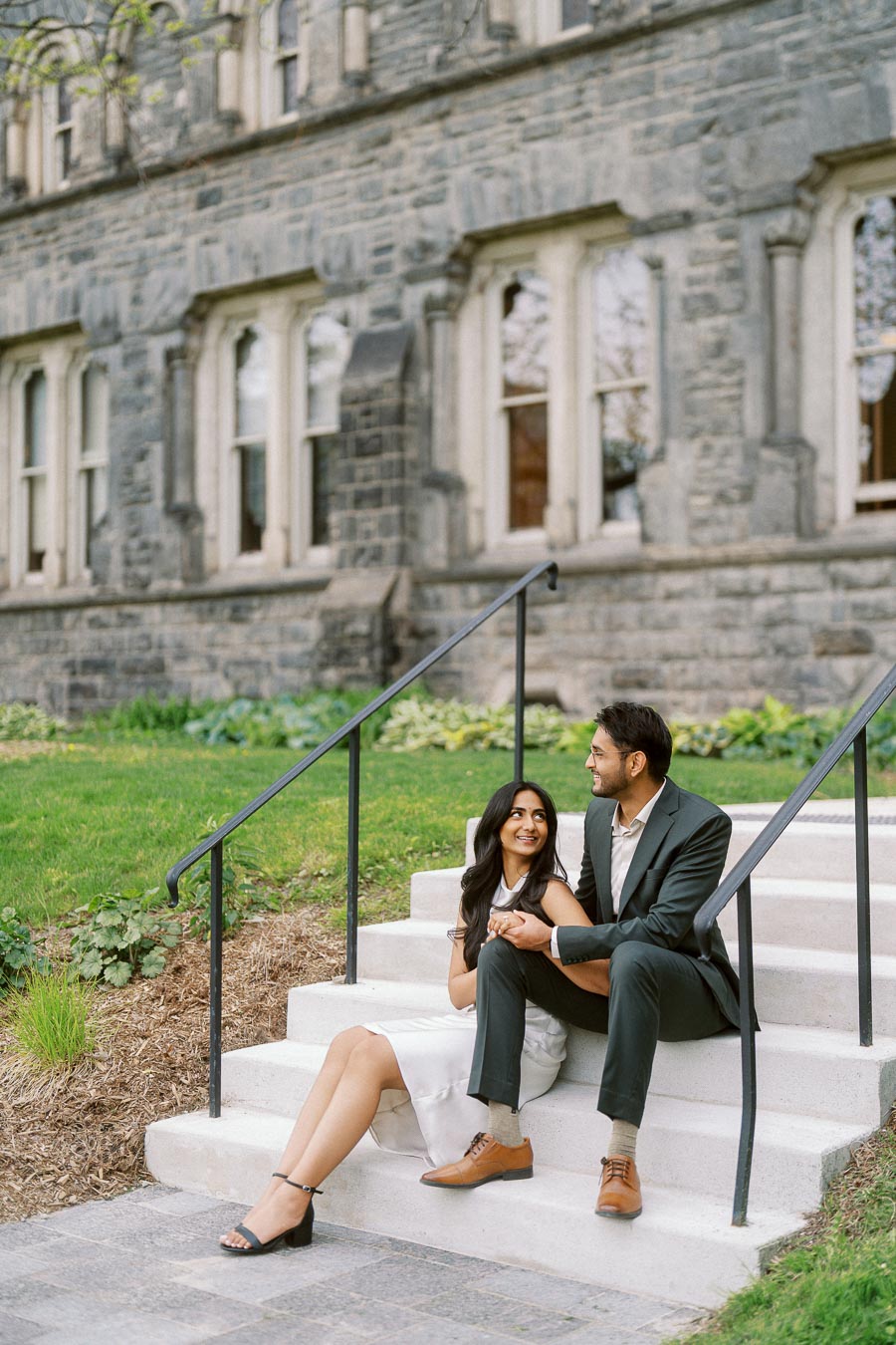 Tej and Priyana sit on outdoor stone steps, holding hands and looking at each other in front of a historic stone building at the University of Toronto.