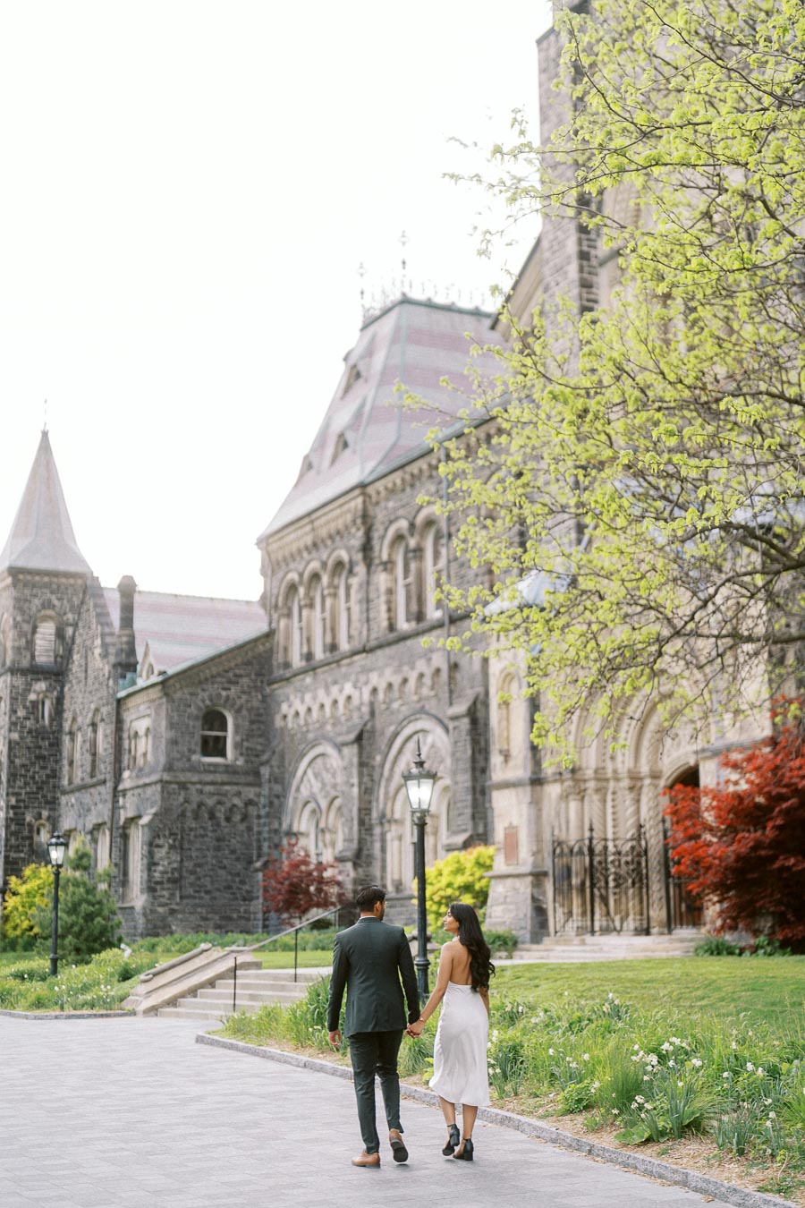 A couple holding hands and walking along a paved path in front of historic stone buildings and spring trees at the University of Toronto.