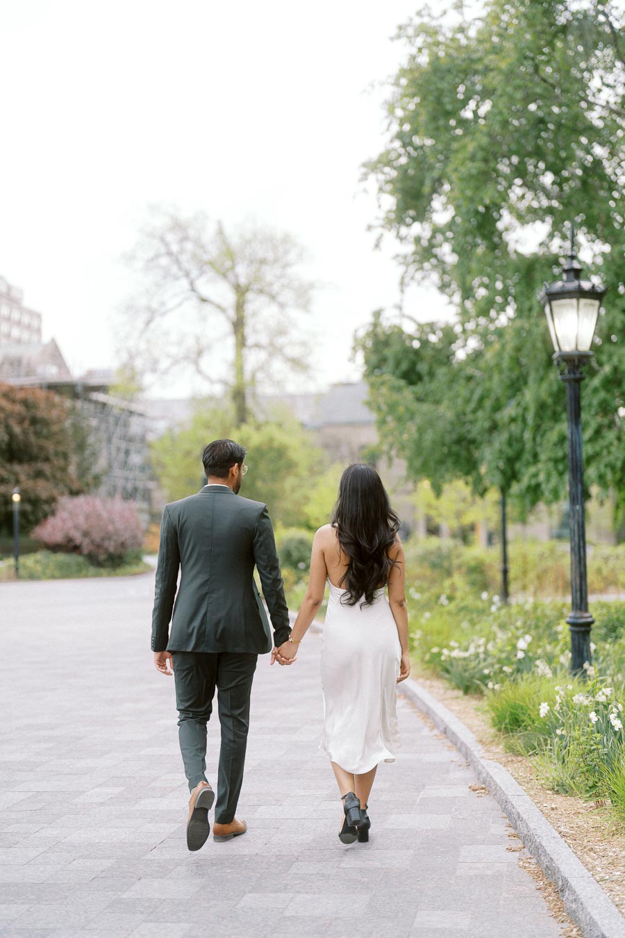 A couple walks hand in hand down a paved campus path at the University of Toronto, surrounded by trees and greenery.
