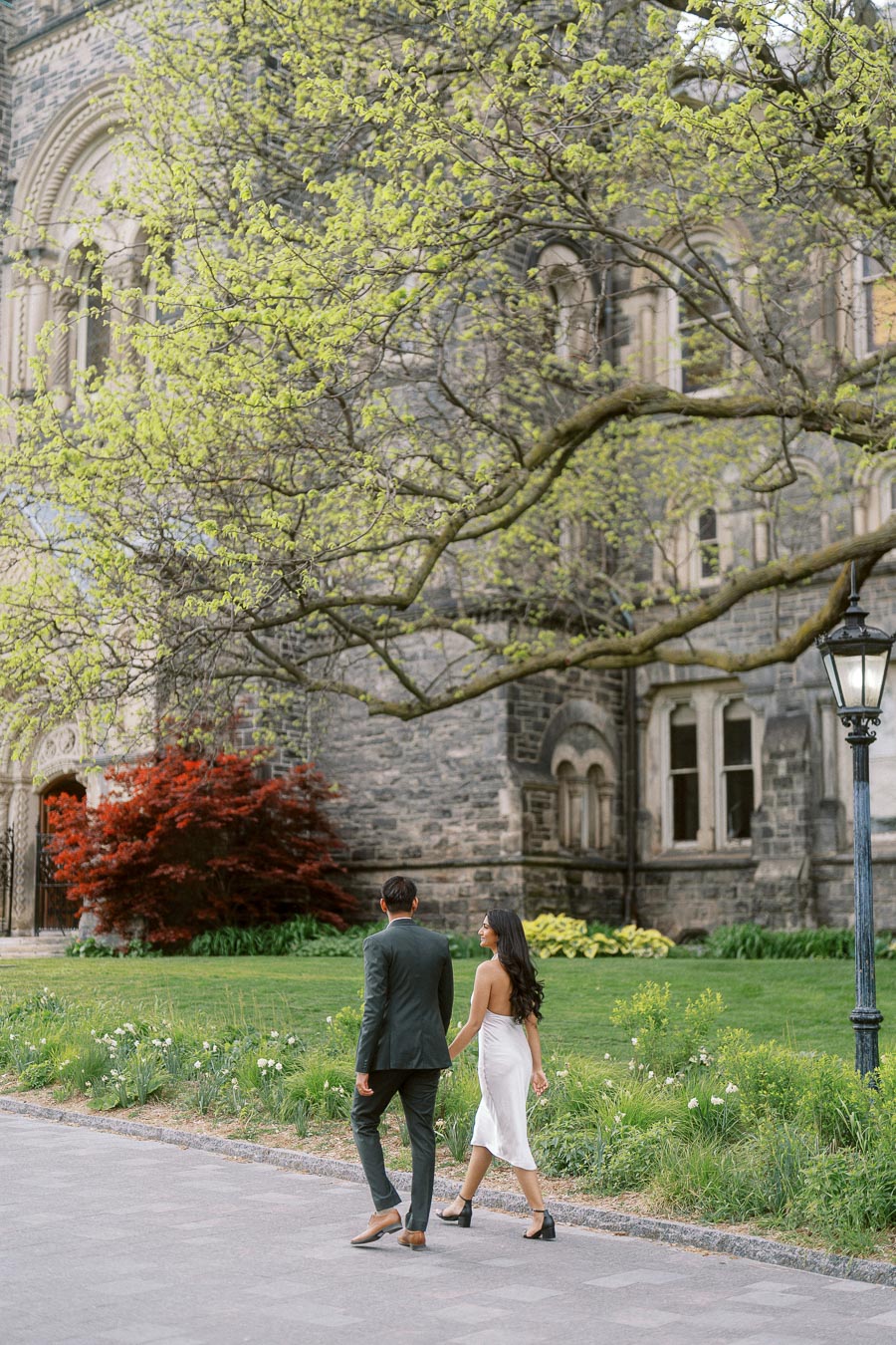 A couple walks together on a campus path with historic stone buildings and spring trees at the University of Toronto.