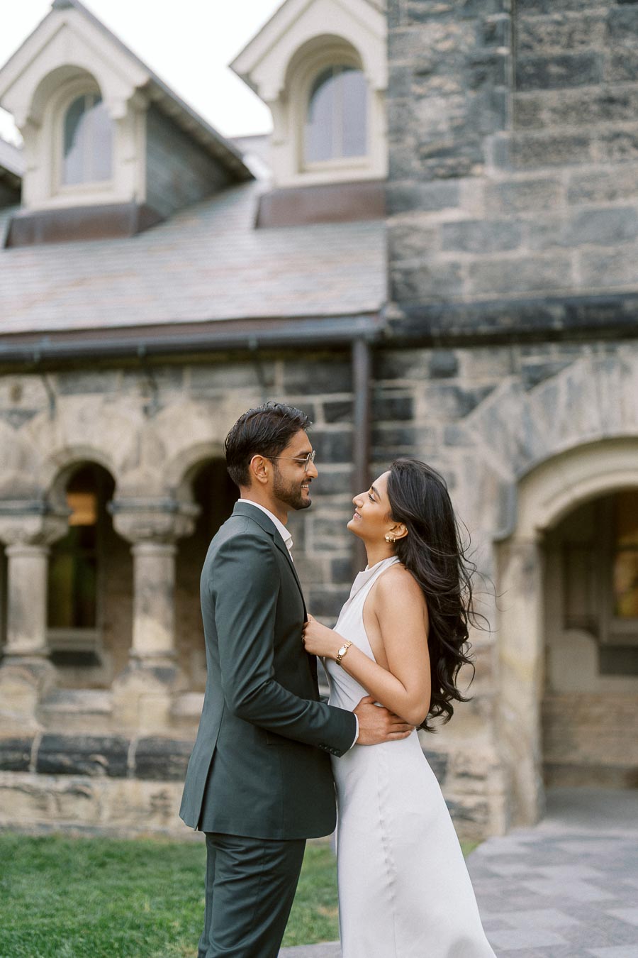 A couple, Tej and Priyana, stand close together smiling in front of a historic stone building at the University of Toronto.