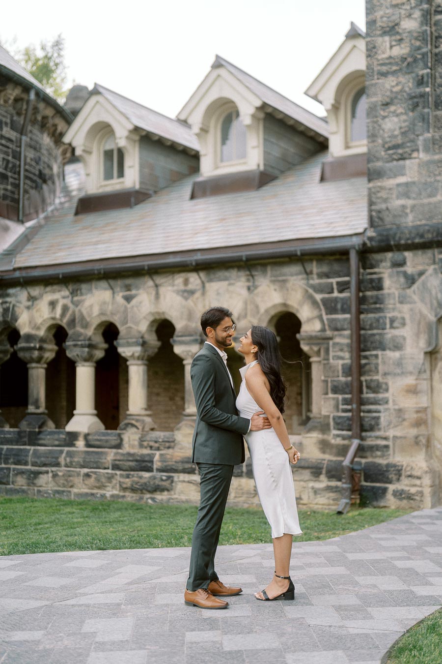 Tej and Priyana stand facing each other on a stone path, with historic stone arches of the University of Toronto in the background.