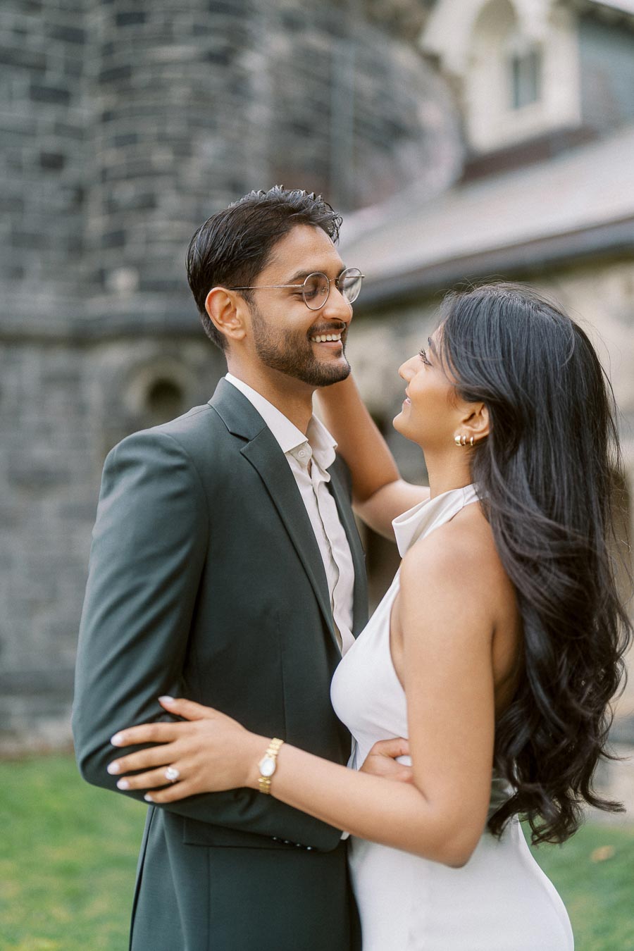 A couple embracing and smiling at each other during an engagement photoshoot, standing in front of historic stone architecture at University of Toronto.