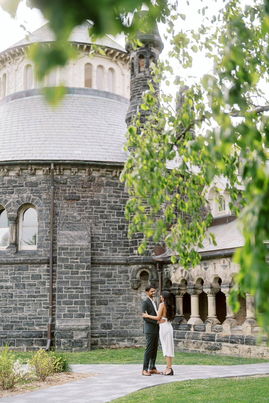 A couple stands close together on a pathway in front of a historic stone building at the University of Toronto, partially framed by green tree branches.