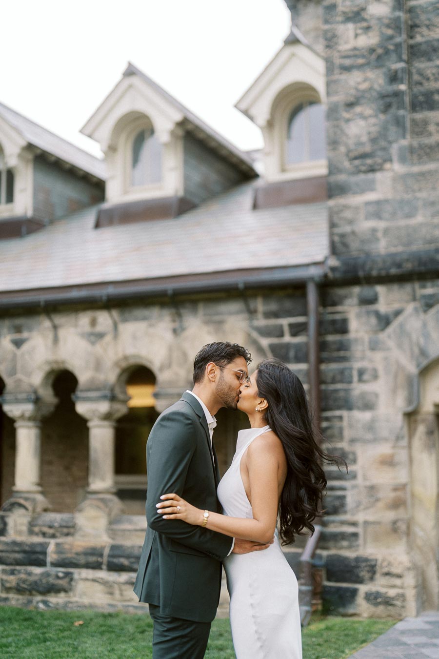 A couple, Tej and Priyana, kiss in front of a historic stone building at the University of Toronto St. George campus.