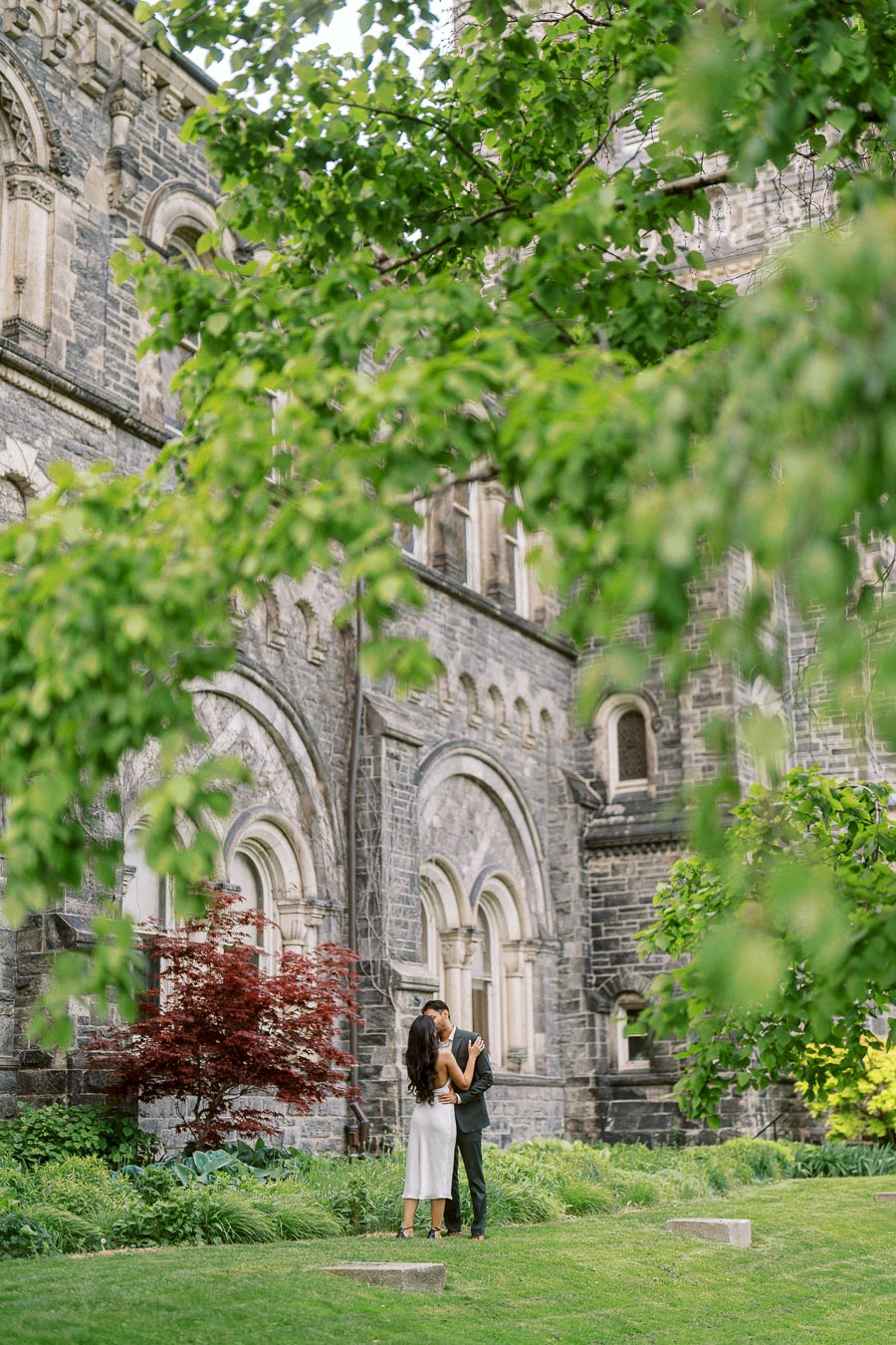 A couple stands embracing on a green lawn in front of a historic stone building at the University of Toronto, framed by leafy branches.