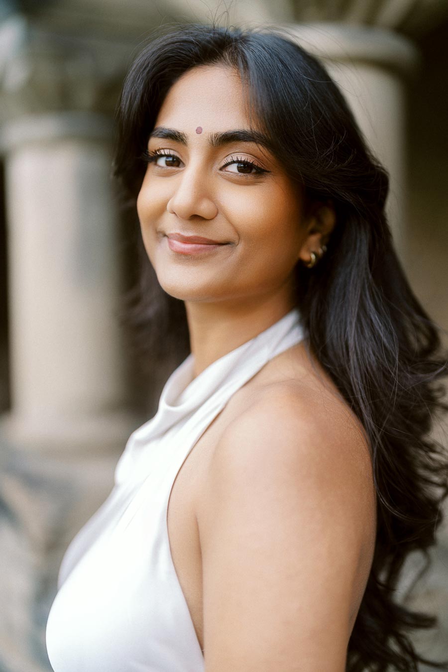 Close-up portrait of Priyana in a white dress, standing outdoors with stone columns in the background at University of Toronto.