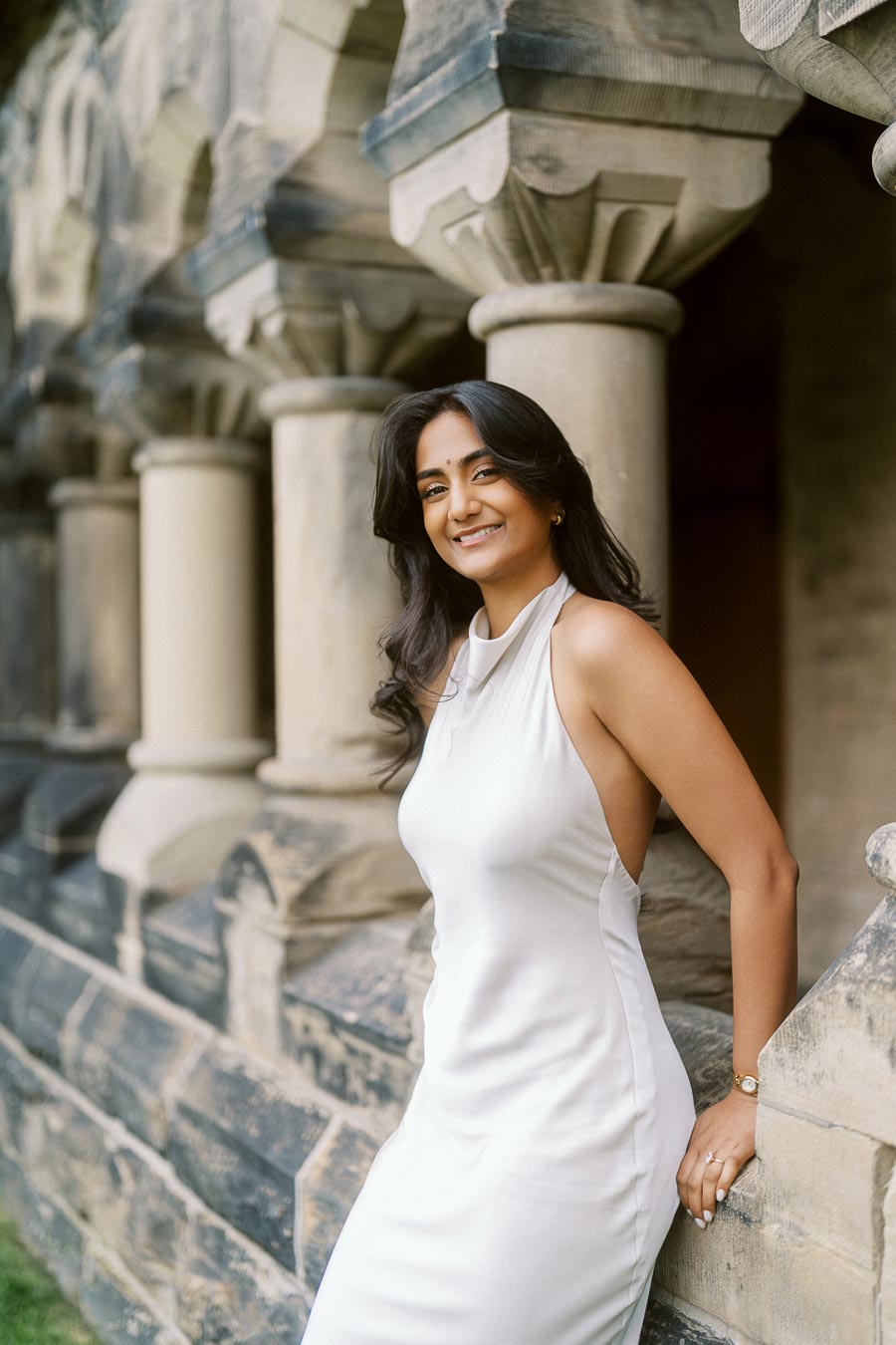 A woman in a white dress stands by historic stone columns at the University of Toronto St. George campus, smiling at the camera.