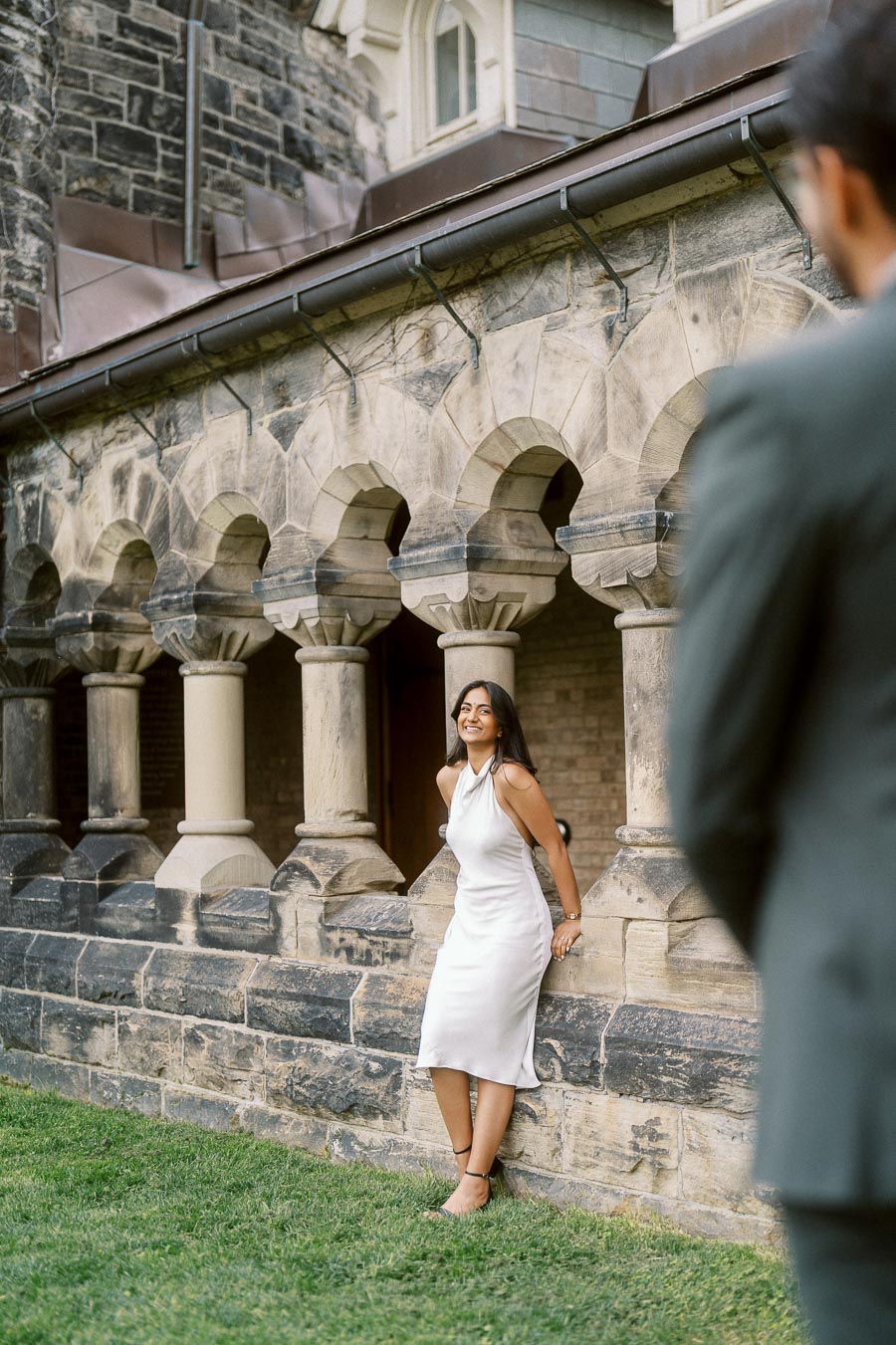 Priyana poses in a white dress against a historic stone archway at the University of Toronto while Tej stands in the foreground.