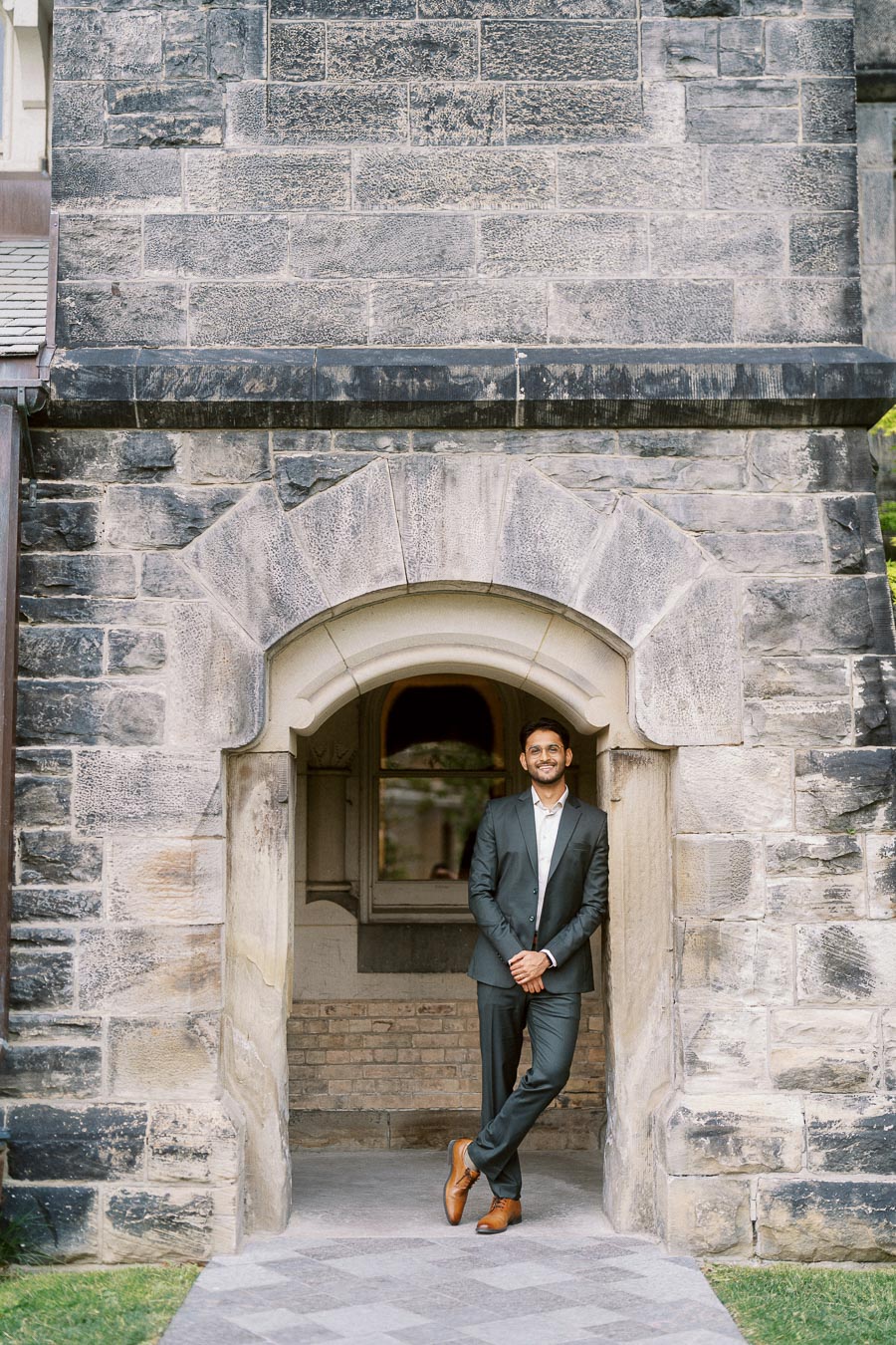 Tej stands in a dark suit, leaning casually against a stone archway at the University of Toronto campus.