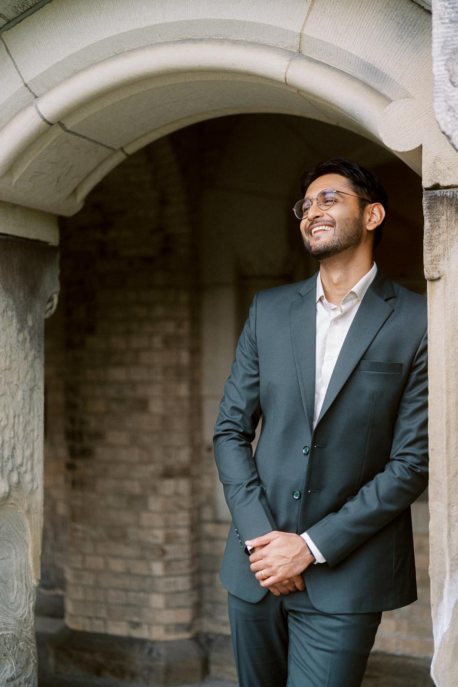 A man in a dark suit leans against a stone archway at the University of Toronto, smiling softly with hands folded.