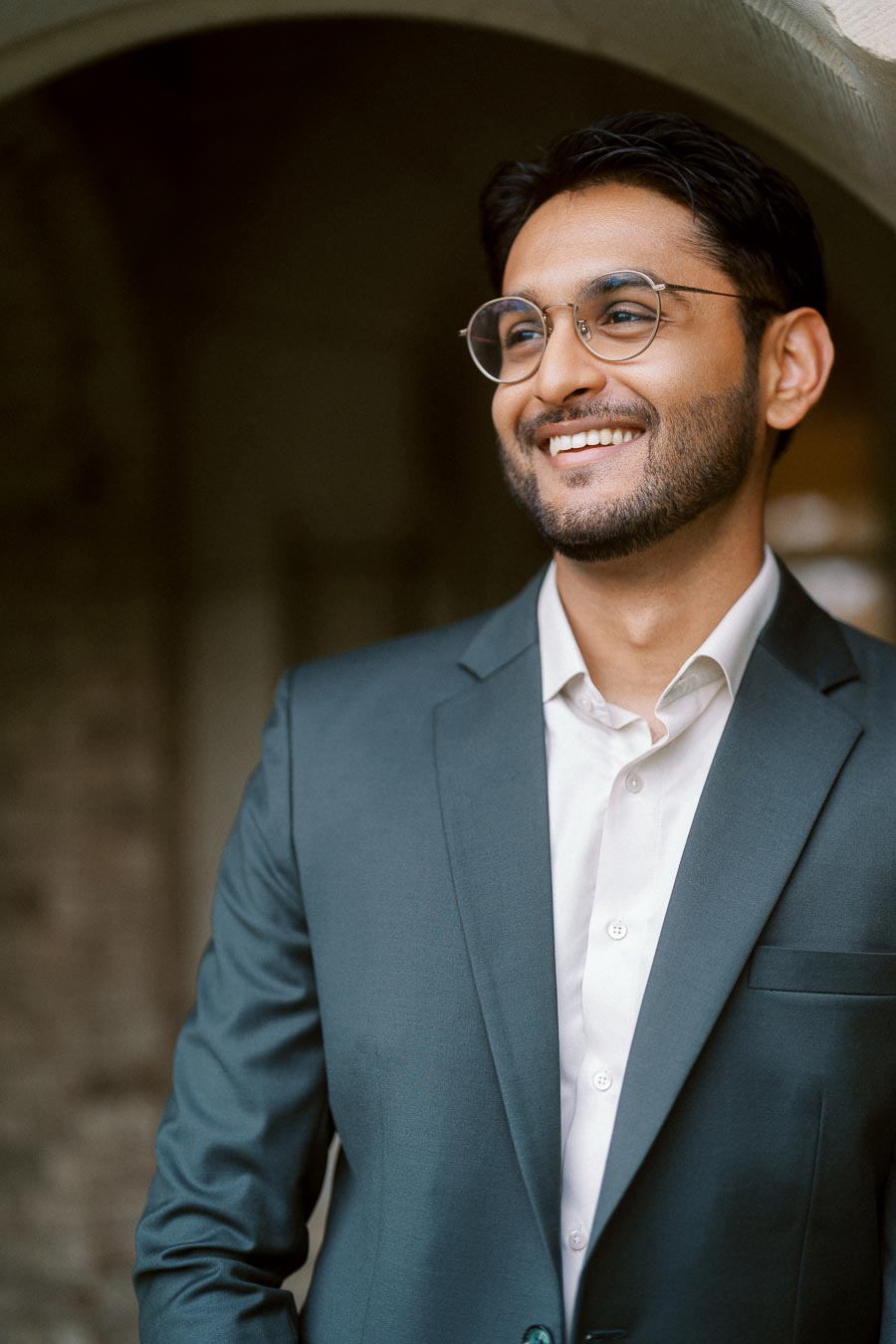 A man in a suit and glasses smiles while standing under a stone archway at the University of Toronto campus.