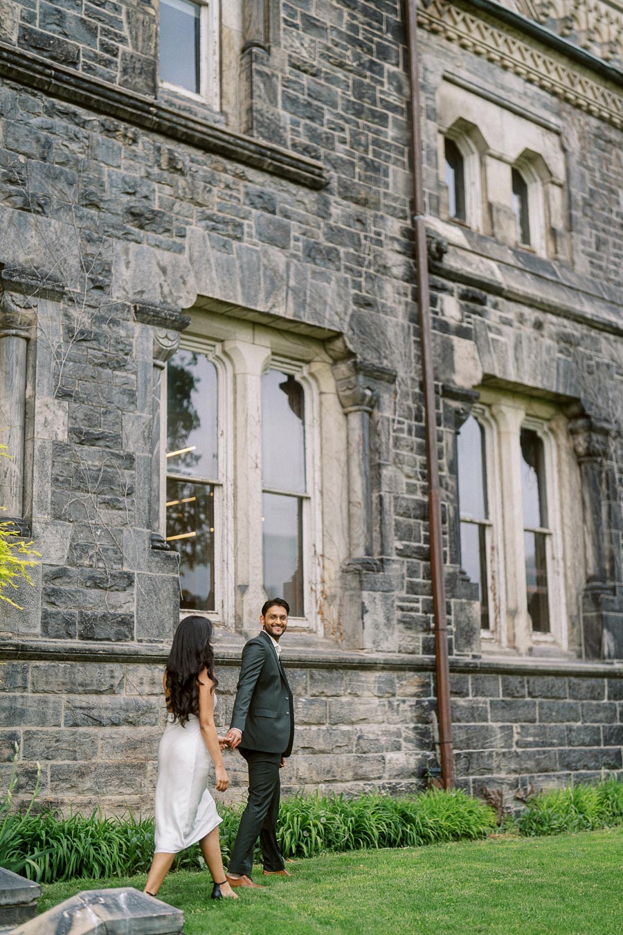 A couple walks hand in hand on a green lawn beside a large stone building at the University of Toronto.